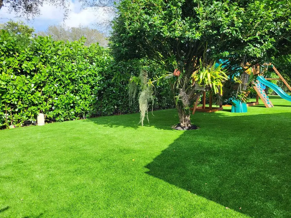 A lush green backyard with a swing set and slide resembling a playground. The lawn, featuring synthetic turf, is bright and well-maintained, bordered by thick green hedges. A tree stands in the center, adorned with hanging plants on this bright, sunny day with some clouds.