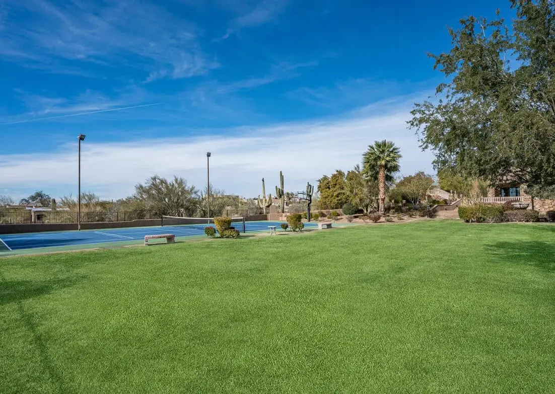 A lush green artificial turf lawn stretches in the foreground, leading to a blue tennis court on the left. The background features desert plants, including a palm tree and cacti, under a clear blue sky.