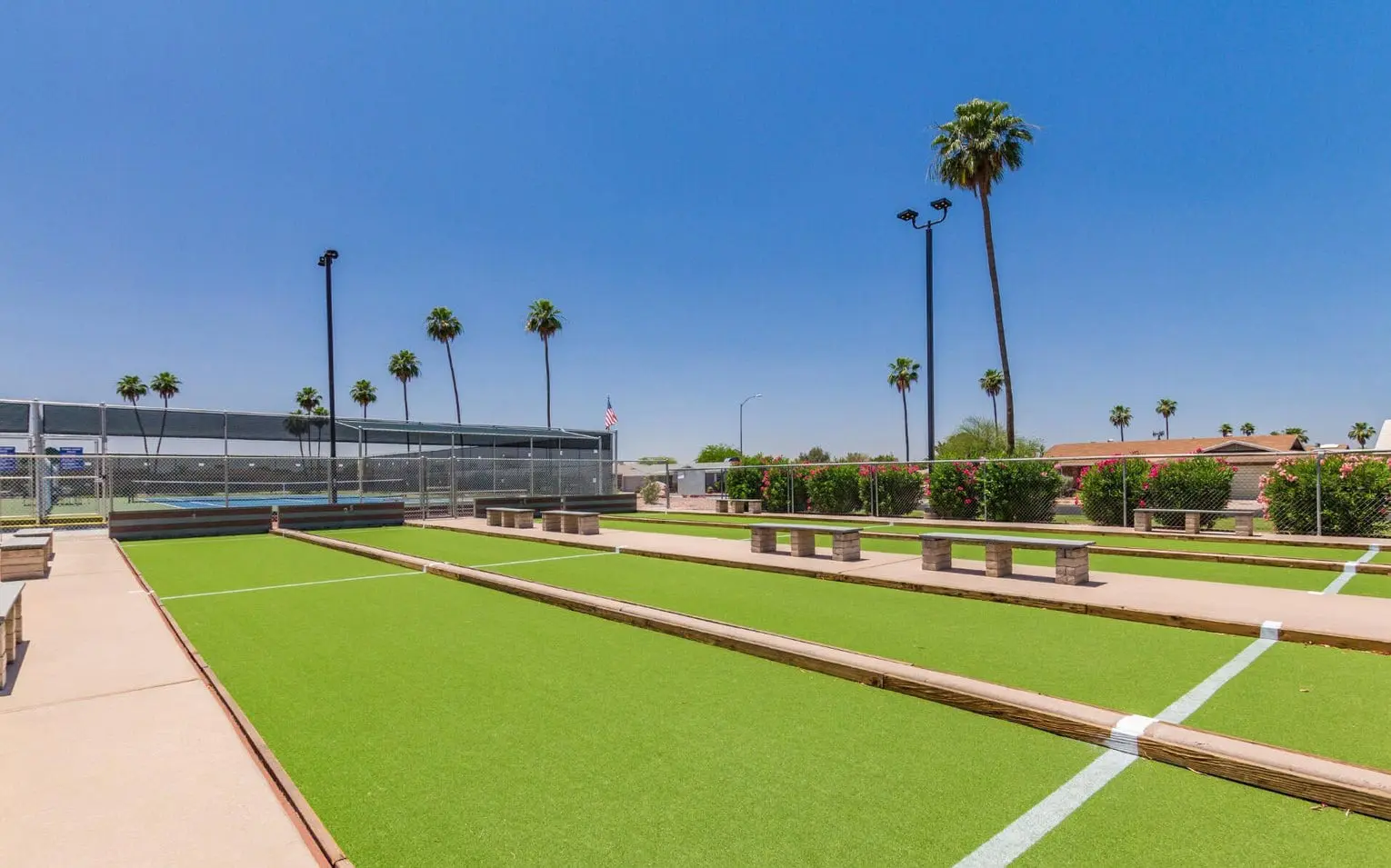 Outdoor bocce ball courts with lush green synthetic turf, bordered by wooden planks. Palm trees and a clear blue sky enhance the scenic view, while benches line the sides. A mesh fence adds character on one side, making it a picturesque spot for enjoying sports fields in style.