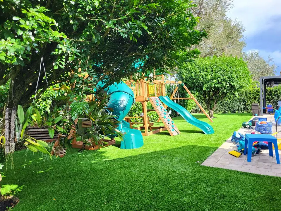 A backyard with vibrant green grass, possibly from synthetic turf installations, features a wooden playset. The playset includes a green spiral slide, another green slide, and climbing elements. Trees and bushes surround the area, while scattered toys and a blue table are visible.