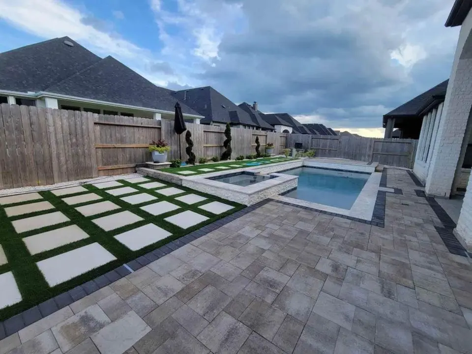 A backyard features a modern swimming pool with a connected hot tub. The outdoor space is enhanced by paver installation and includes neatly trimmed grass squares. A wooden fence separates the yard from neighboring houses under a partly cloudy sky.