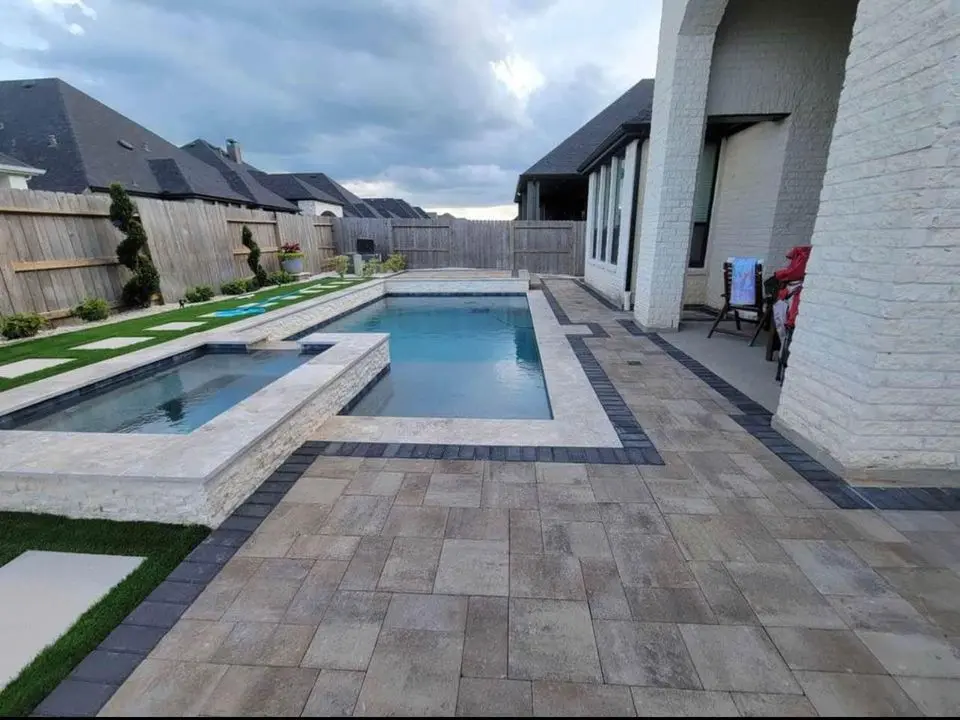 A modern backyard showcases quality craftsmanship with a rectangular swimming pool and an adjacent hot tub, both elegantly framed by light and dark paver tiles. The well-designed outdoor space is enhanced by a row of grass and small trees lining the fenced yard, alongside a white brick house under cloudy skies.