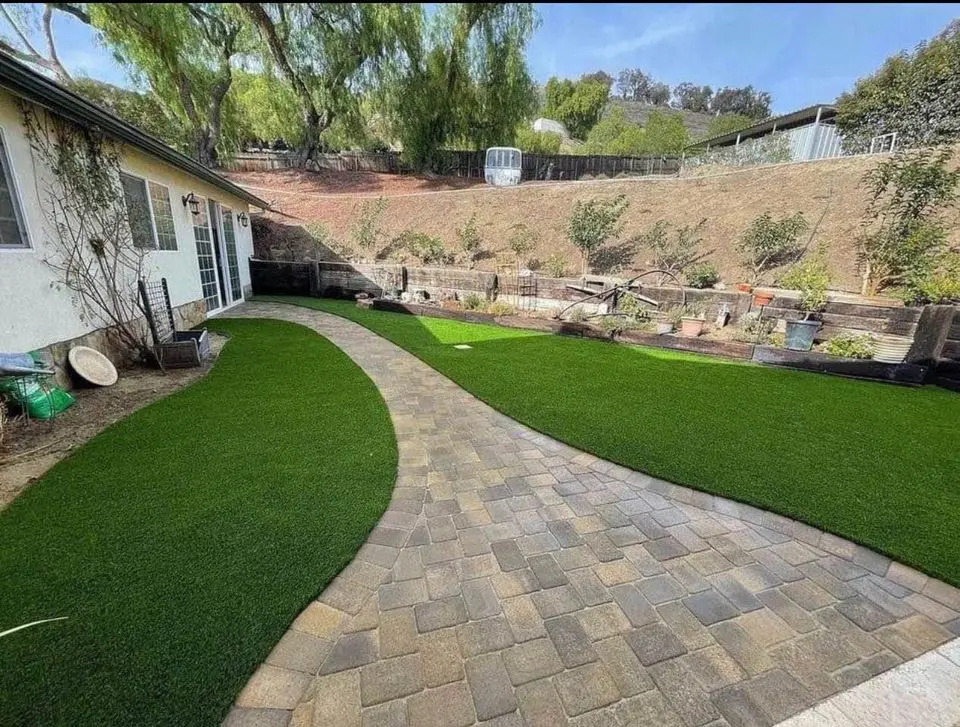 A backyard with a curving stone path dividing two areas of neatly trimmed artificial grass, showcasing the expertise of a skilled artificial grass installer. The yard is bordered by a gentle slope with trees and potted plants, while a white house stands on the left under the clear blue sky.