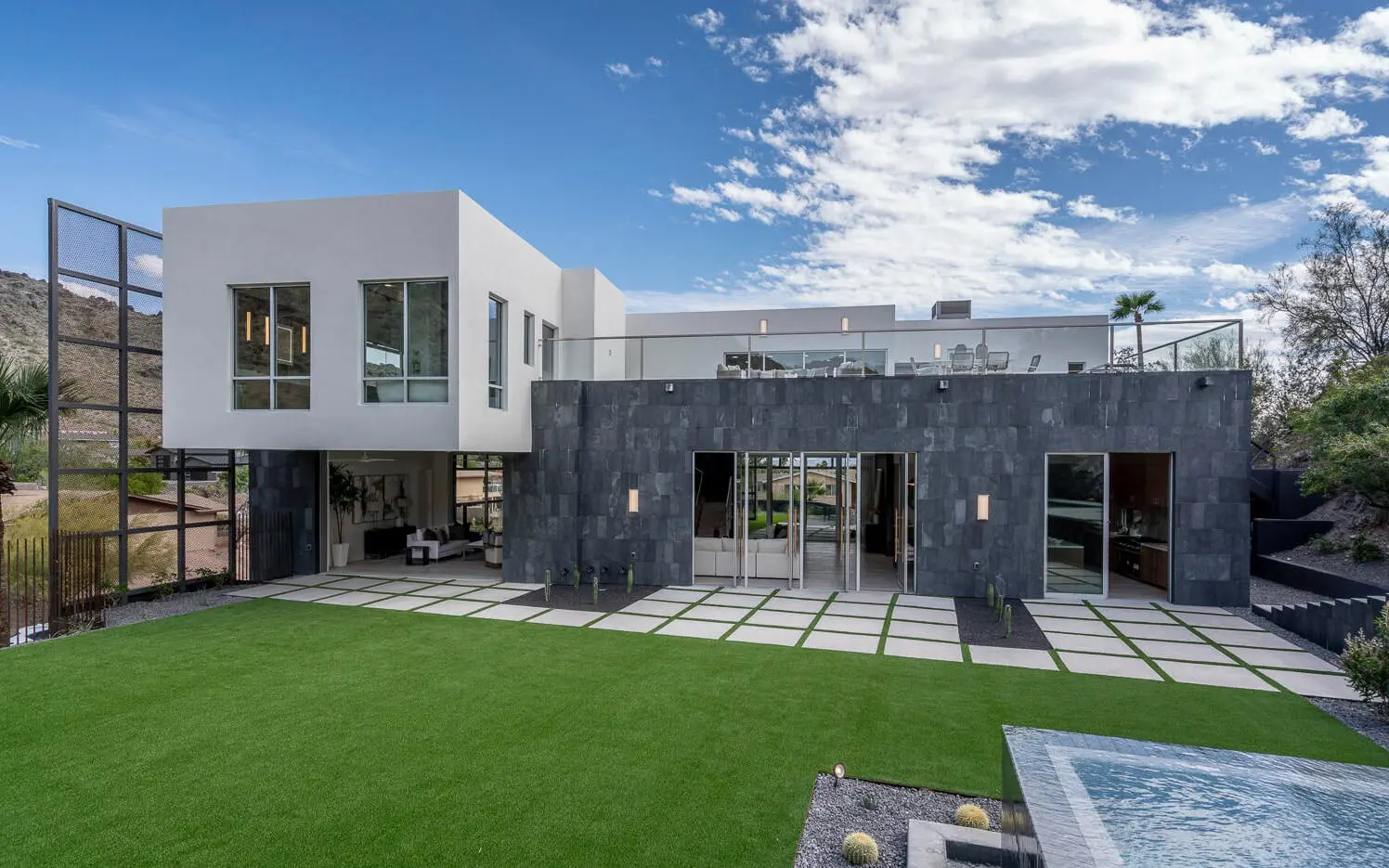 Modern two-story house with a white and dark gray exterior, featuring large windows and a rooftop terrace. The foreground includes a well-maintained lawn with synthetic turf, a patio area, and a rectangular water feature. The sky is partly cloudy.