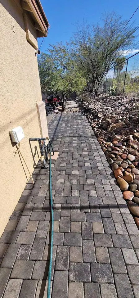 A stone-tiled walkway, a testament to quality craftsmanship, winds next to a beige building with a garden hose along the path. On the right, rocks and trees gracefully line the edge, enhancing the serene outdoor space under a clear blue sky.