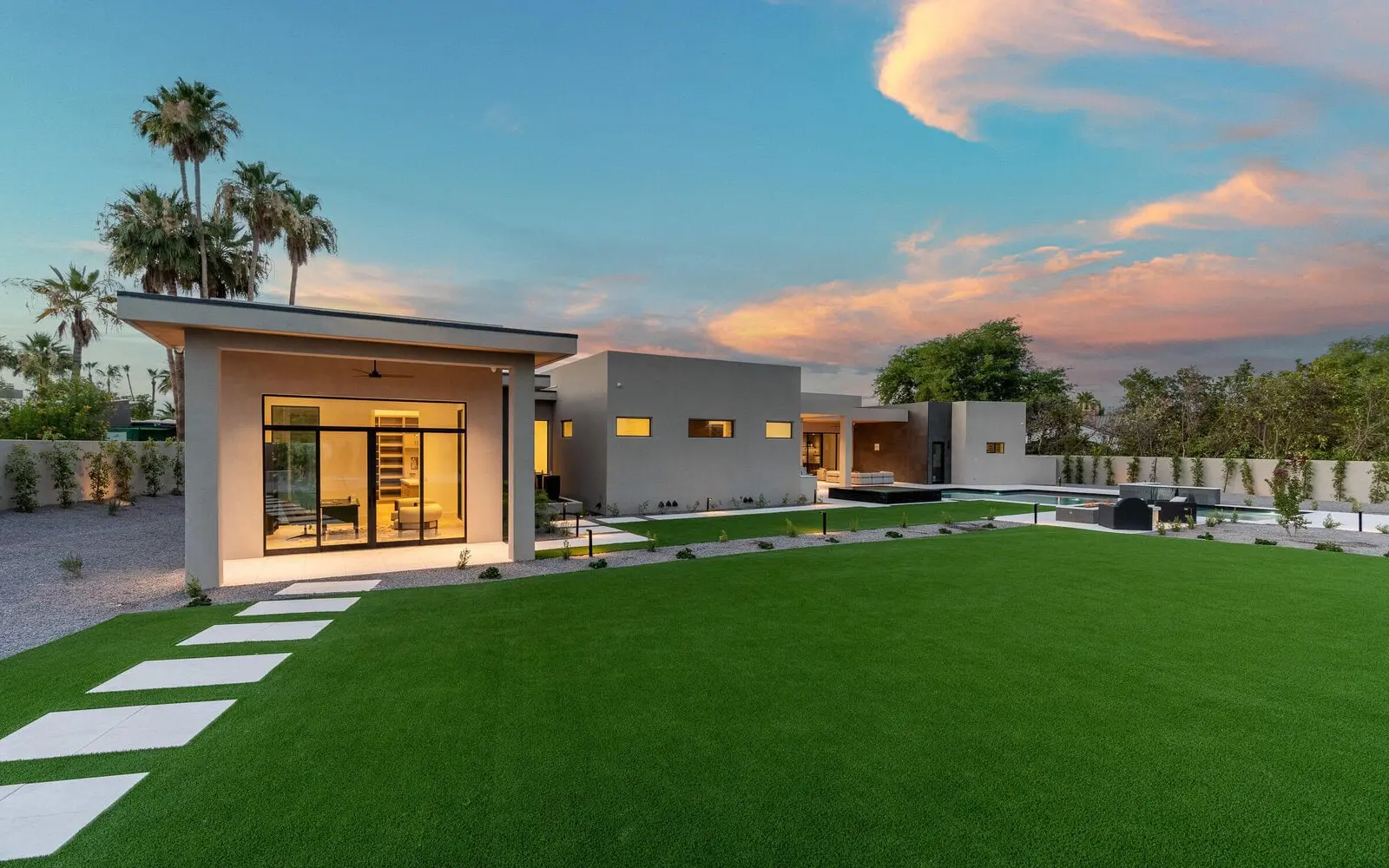 Modern house with large glass windows, surrounded by a spacious green lawn of synthetic turf and stepping stones. The sky at sunset adds vibrant colors, with palm trees in the background. The architecture features clean lines and a minimalist design. Contact for free quote on artificial grass installation.