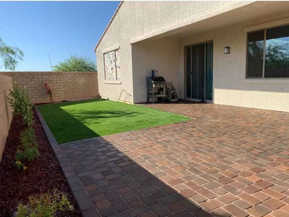 A backyard boasts a patch of green grass, surrounded by brick paving. An artificial grass installer offers lush alternatives nearby. A grill sits near sliding glass doors, enclosed by a light-colored fence, with plants along the edges and sunlight casting shadows on the ground.