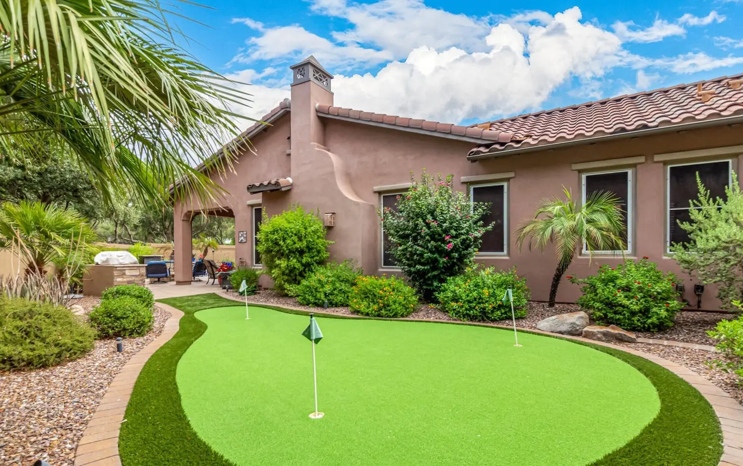 A backyard featuring custom putting greens, perfect for year-round golfing, is framed by lush shrubs and palm trees. The house boasts a stucco exterior and tiled roof, with a barbecue and seating area visible under the partly cloudy Phoenix sky.