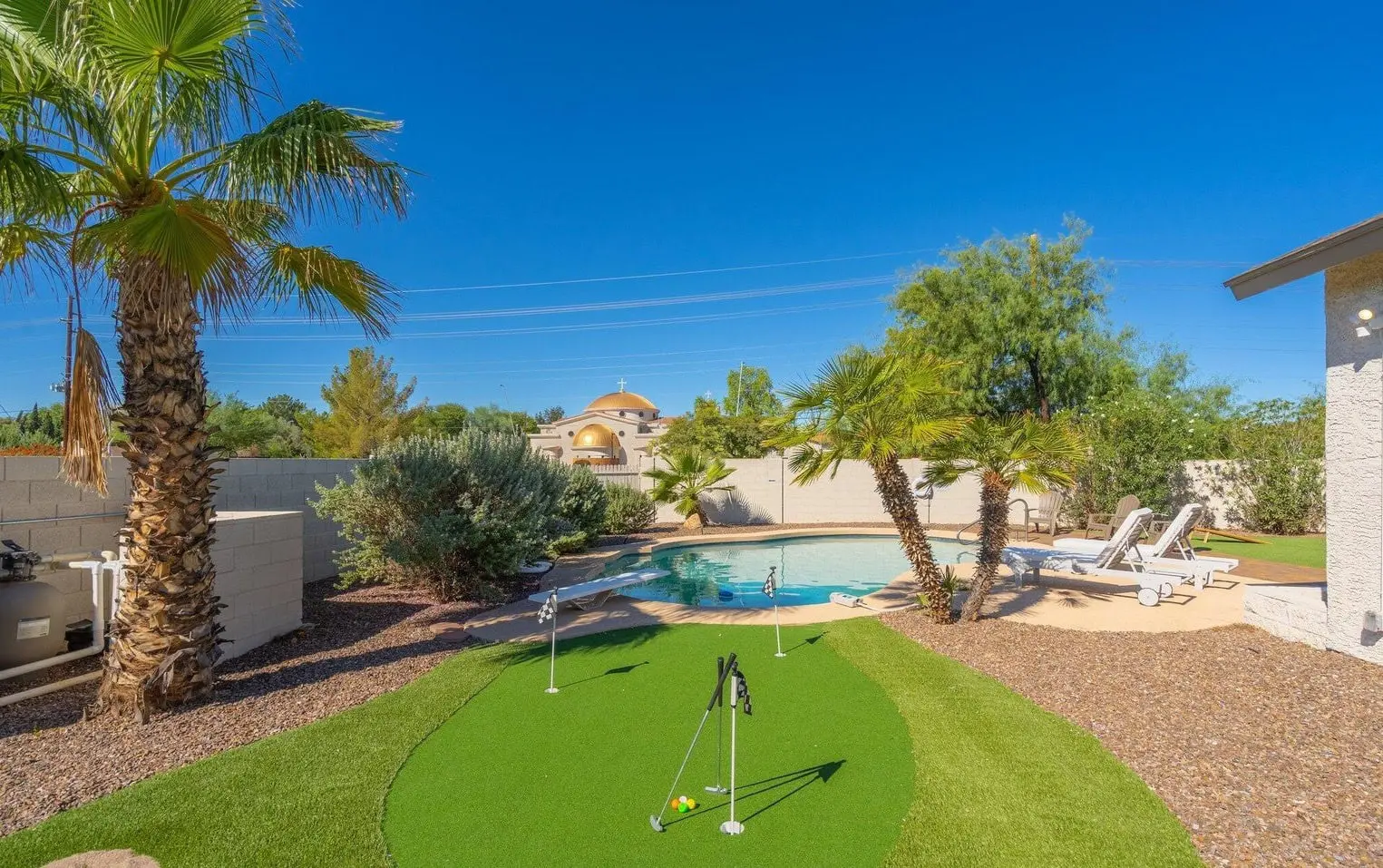 Image of a sunny Phoenix backyard featuring a palm tree, backyard putting greens with flags, and a swimming pool surrounded by lounge chairs. The area is enclosed with a block wall, and there are shrubs and a clear blue sky in the background, perfect for year-round golfing enjoyment.