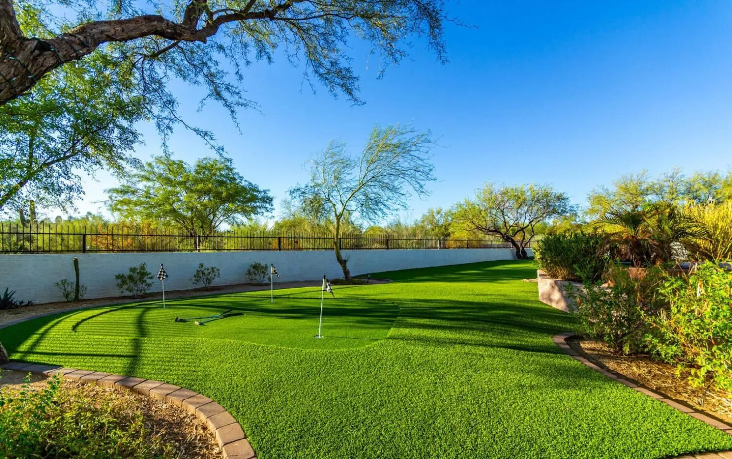 Nestled in the heart of Glendale AZ, this lush backyard boasts an outdoor space with a green putting course featuring flag-marked holes. Surrounded by desert plants and trees under a bright blue sky, the area benefits from artificial grass installation and is enclosed by a white fence.