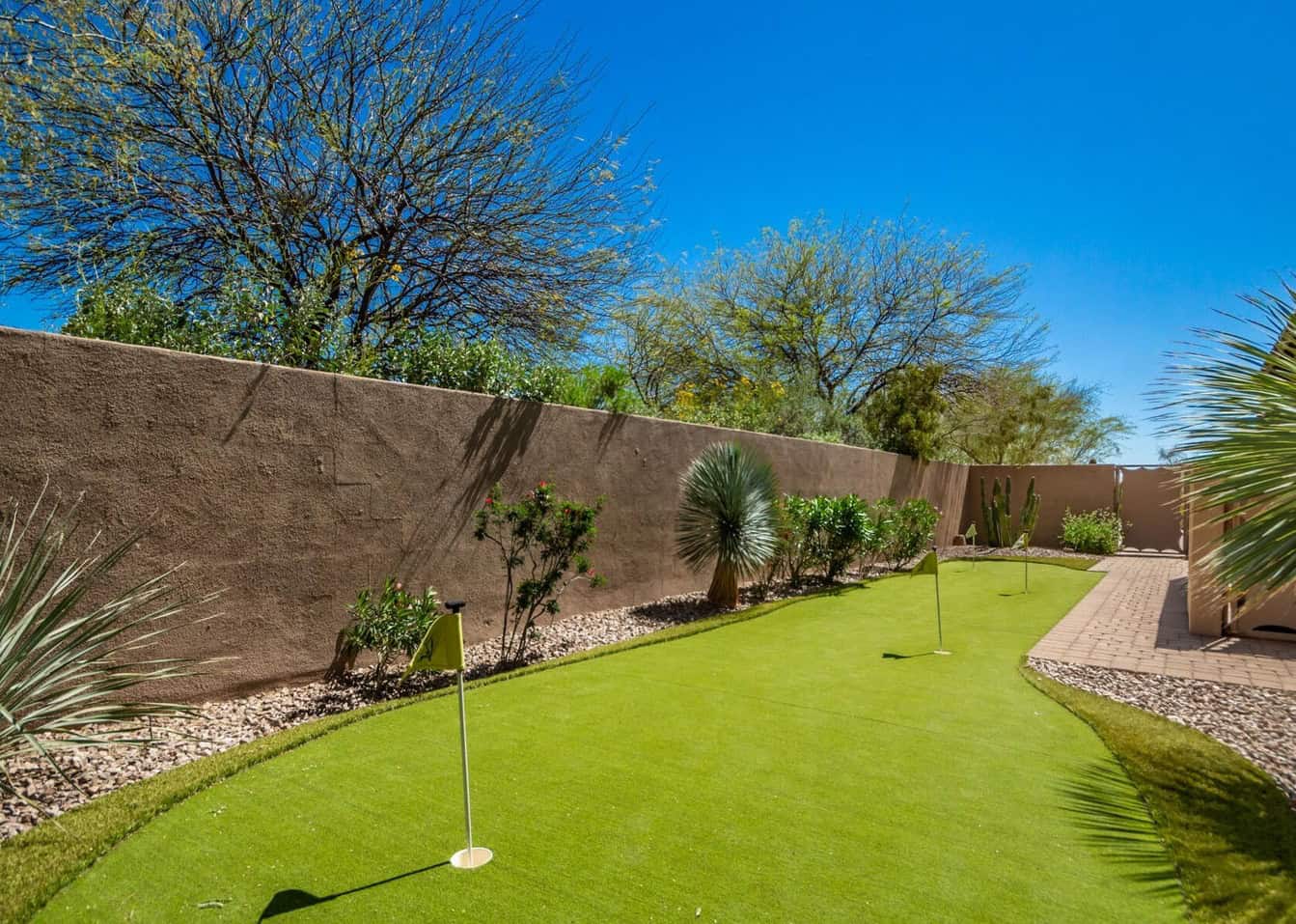 A Phoenix backyard retreat showcases a small putting green with two golf holes, bordered by lush plants and shrubs. A stone pathway graces the right side, set against a backdrop of clear blue sky and trees, making it perfect for year-round golfing pleasure.