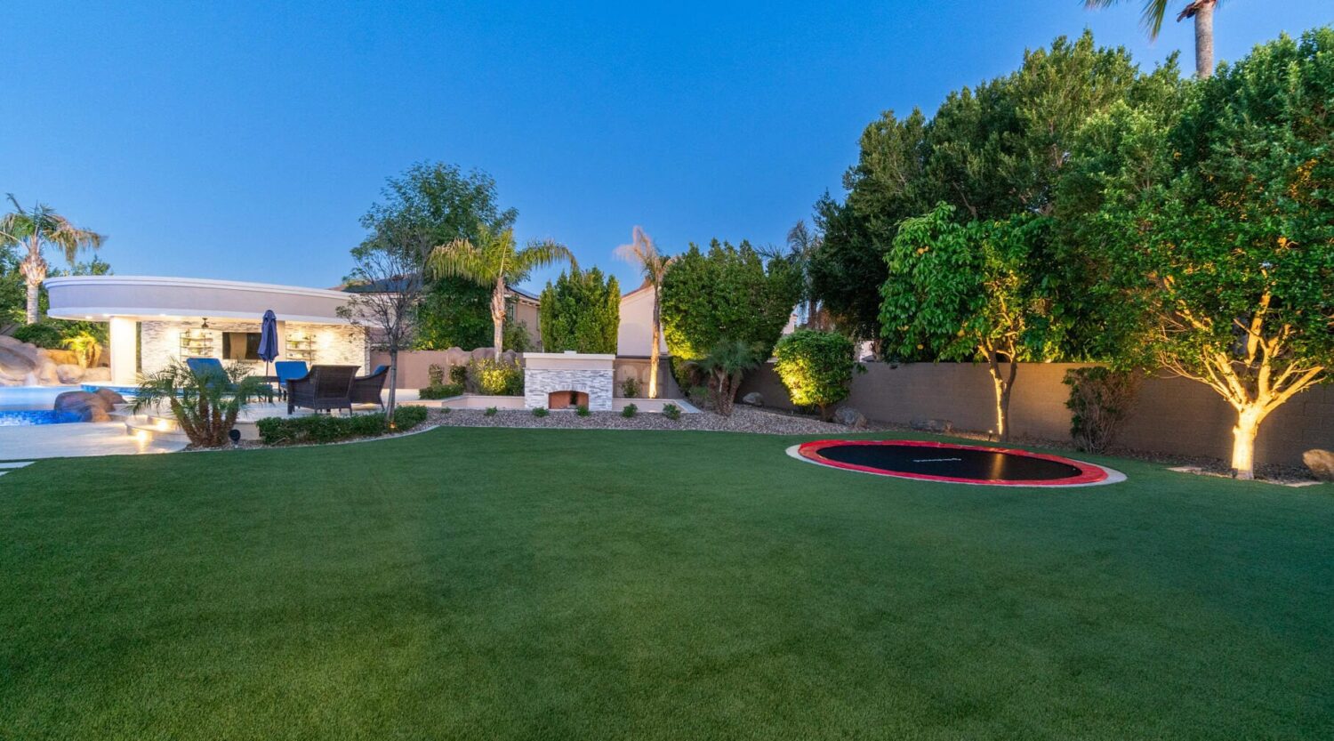 Spacious backyard with a lush, pet-friendly lawn featuring a sunken trampoline. Trees surround the area, adding privacy. A patio with a seating area and a pool with a waterfall are visible under the clear blue sky.