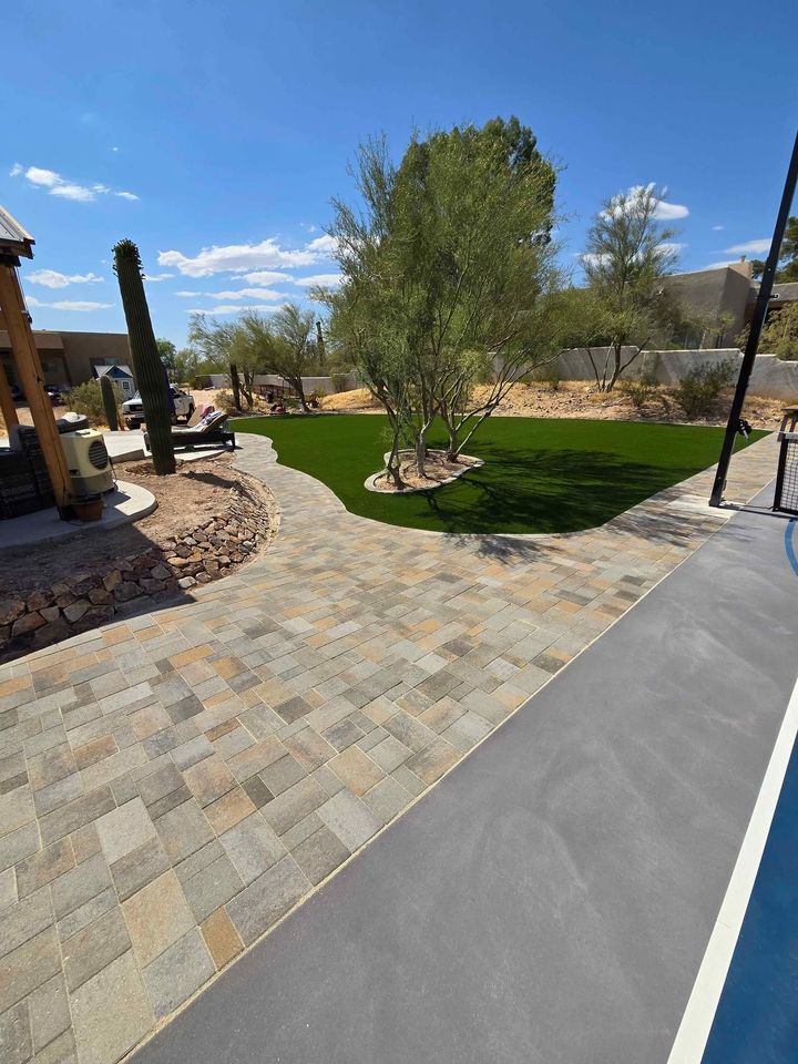 A backyard featuring a stone patio, lush synthetic turf installations, and desert landscaping with cacti and trees. Set under a clear blue sky with scattered clouds, a swimming pool edge is visible in the foreground. Perfect for those seeking an artificial grass installer or a free quote.