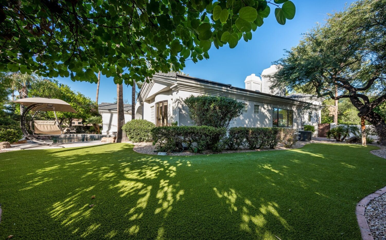 A tranquil backyard with lush artificial turf, trees, and shrubs under a clear blue sky. A shaded seating area with a swing is visible in the corner, near a swimming pool surrounded by plants, offering a pet-friendly lawn perfect for relaxation.