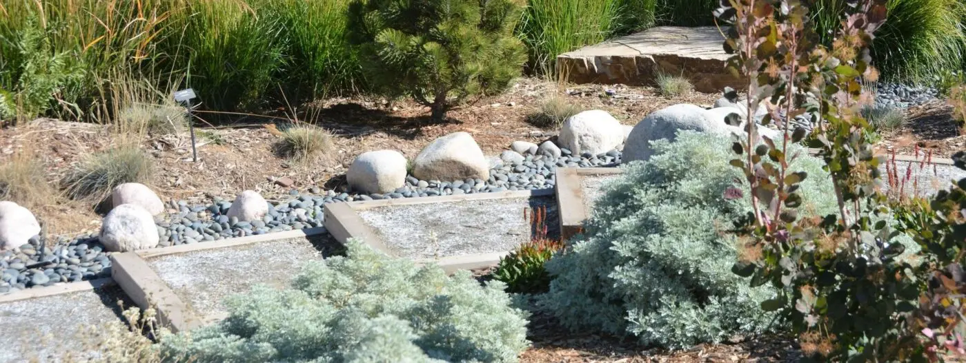 A serene garden scene featuring stone pathways, various shrubs, and large white rocks. The lush greenery is perfectly complemented by synthetic turf installations, creating a harmonious and tranquil landscape design. Contact us today for a free quote on your ideal outdoor transformation.