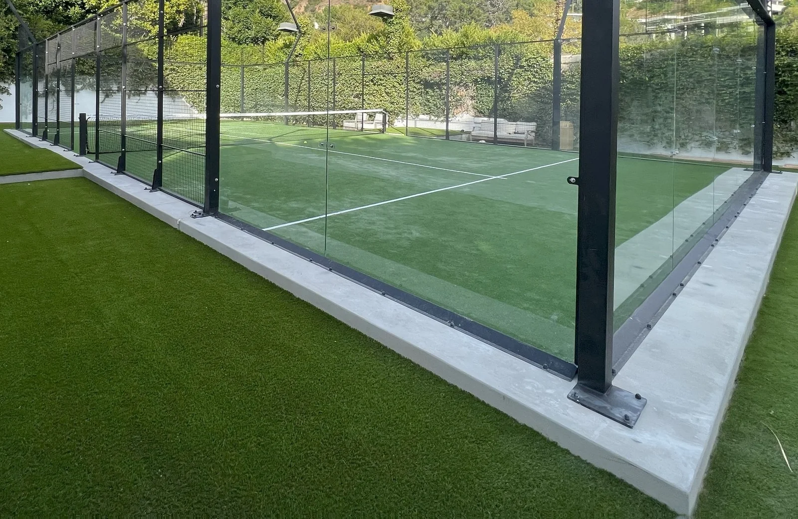 A modern outdoor padel court with a glass enclosure sits on sleek synthetic turf. The court's net and crisp white lines are clearly visible, framed by lush foliage and a clear blue sky, epitomizing the elegance of Phoenix sports fields.