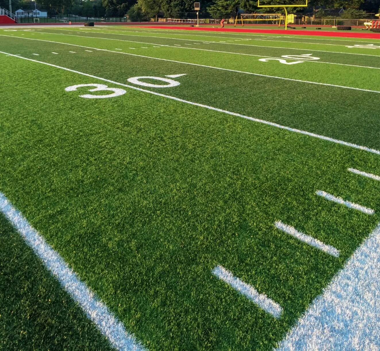 A football field under bright sunlight with a focus on the 30-yard line. The synthetic turf installation is marked with white yard lines, and goalposts are visible in the background. The artificial grass appears well-maintained and vibrant green.