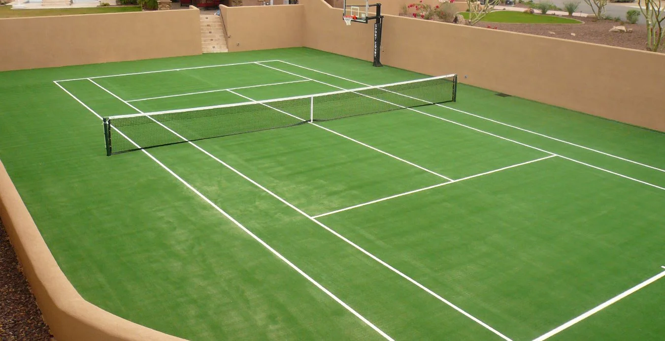 A small tennis court with a green artificial turf surface and white boundary lines is enclosed by tan walls. The court features a net in the center and a basketball hoop against one wall. Steps lead up to a pathway in the background, creating a perfect blend of sports fields.