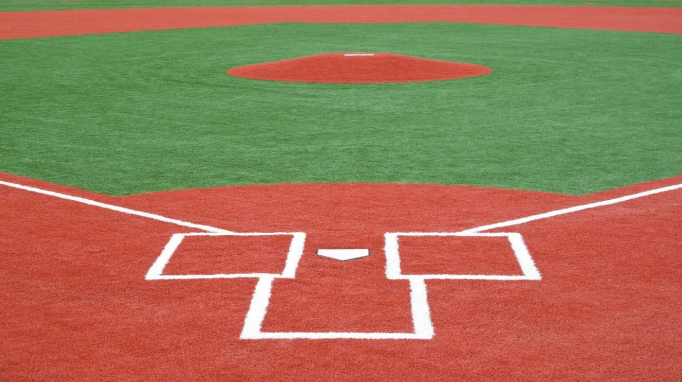 Close-up of a baseball field's home plate area in Phoenix, featuring a neatly marked batter's box and vibrant red clay. The green synthetic turf installations are visible in the background, with a clear path leading up to the pitcher's mound.