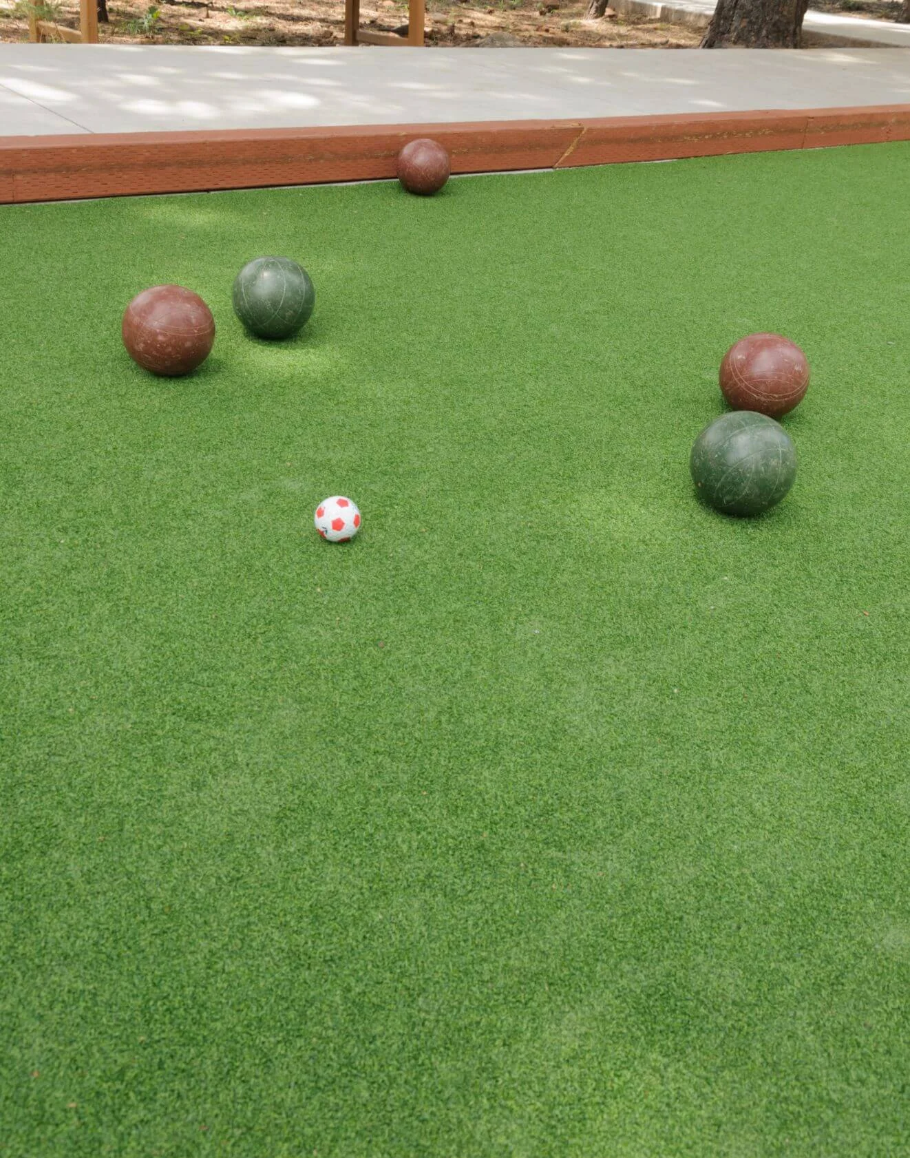 A bocce ball court with four large balls, two brown and two green, scattered around a small white ball with red dots on an artificial turf surface. A wooden border surrounds the court.