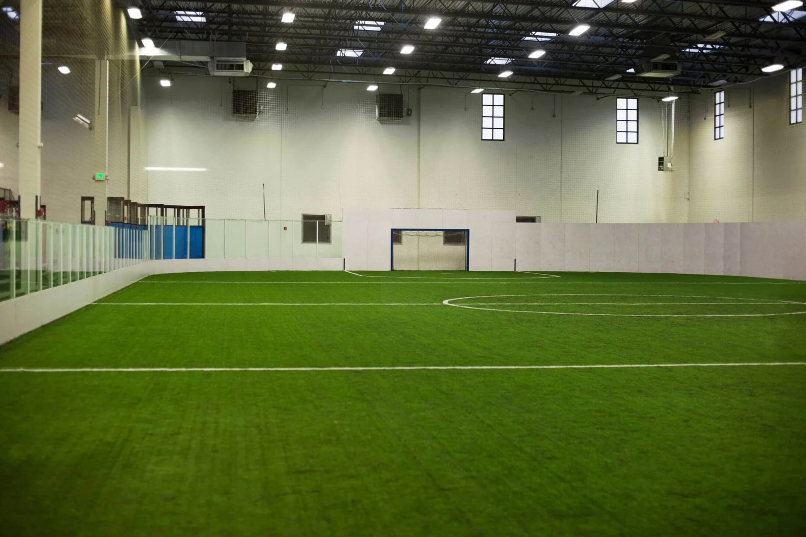 An indoor soccer field in Phoenix, AZ features lush artificial green turf, surrounded by white walls and transparent panels. The high ceiling showcases visible lighting fixtures, and the room stands empty with a goal positioned at one end.