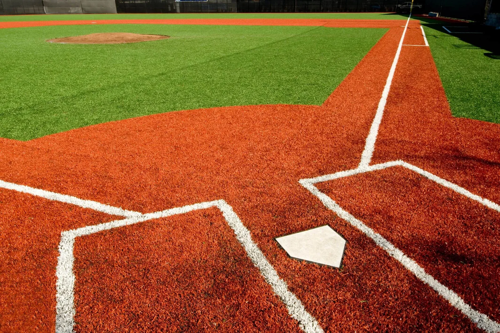 Close-up of a baseball field's home plate area in Phoenix, featuring a neatly marked batter's box and vibrant red clay. The green synthetic turf installations are visible in the background, with a clear path leading up to the pitcher's mound.