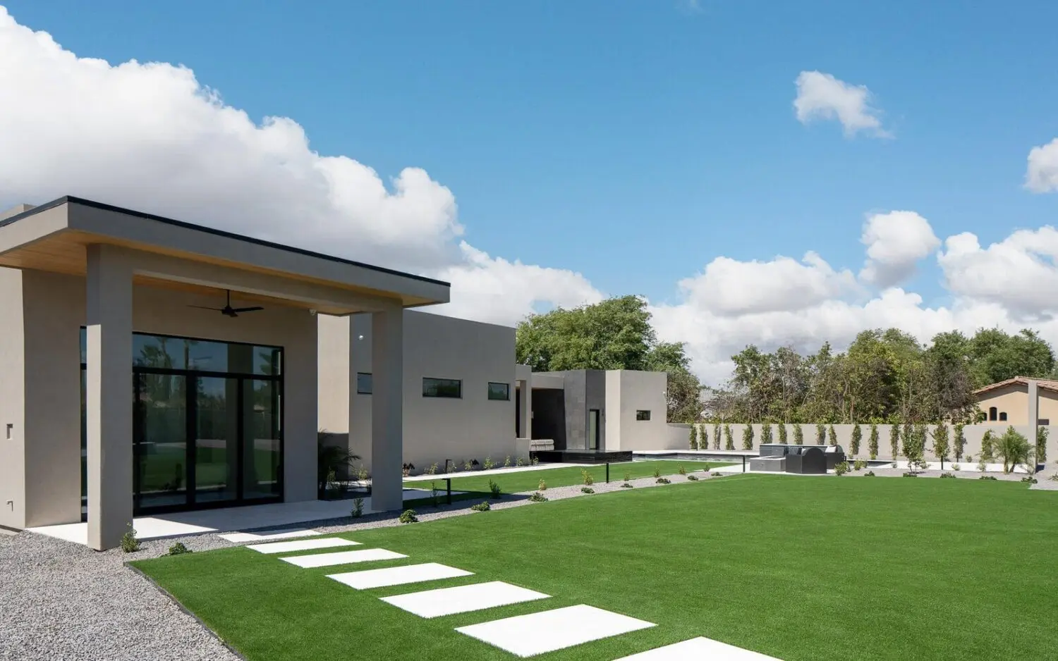 Modern house with large glass doors and beige exterior walls, surrounded by a well-manicured lawn in Glendale, AZ. A stone pathway leads to the entrance. The backyard showcases an impressive outdoor space transformation with trees, bushes, and a paved seating area under a blue sky with clouds.