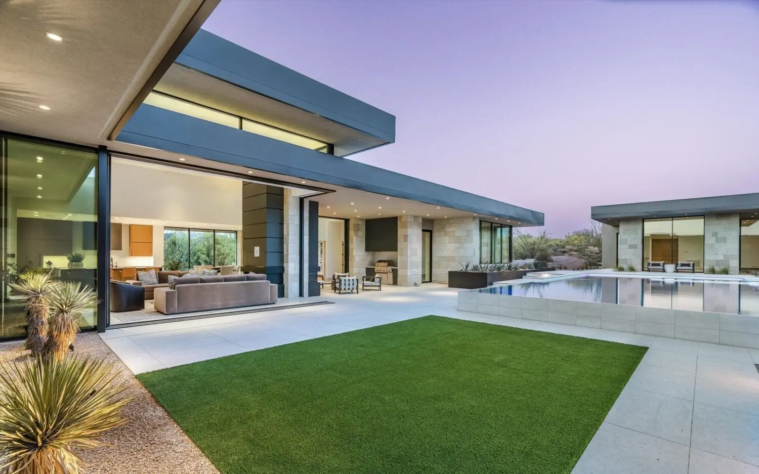 Modern house with large glass windows, an open patio, and a lush green Tempe lawn. The house has a flat roof and minimalist design, surrounded by sparse desert plants. Indoor living space visible through the windows, pool on the right, and a purple dusk sky enhanced by artificial grass.