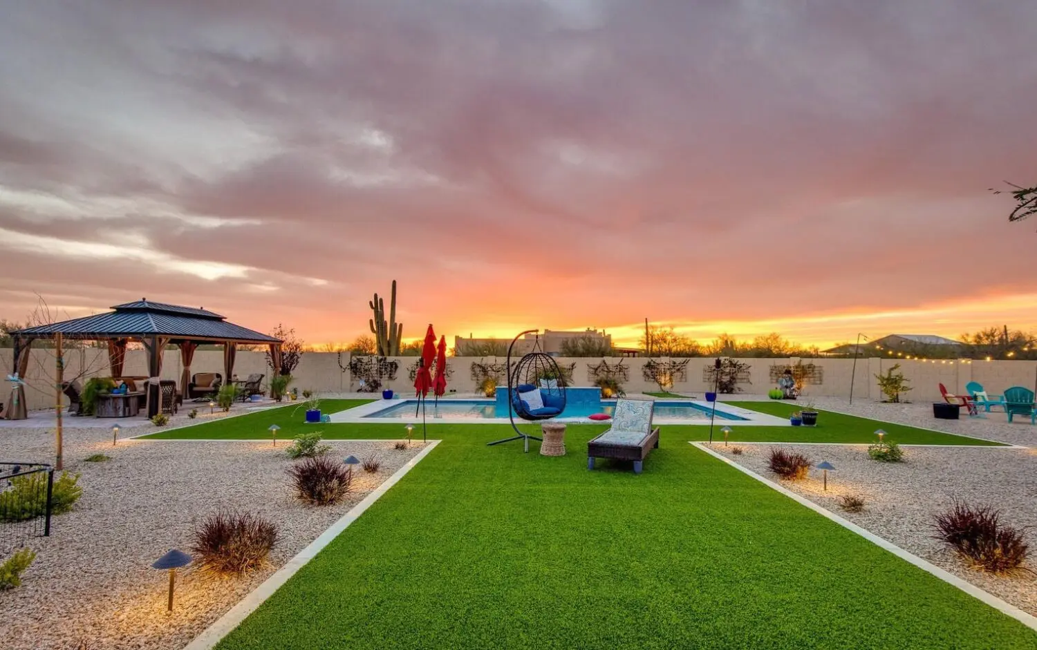 A modern backyard at sunset, featuring a pool, lounge chairs, and a swing chair. The garden is beautifully landscaped with artificial grass and desert plants surrounding the gazebo. The vibrant sky with orange and purple hues reflects off the expertly crafted Tempe lawn.